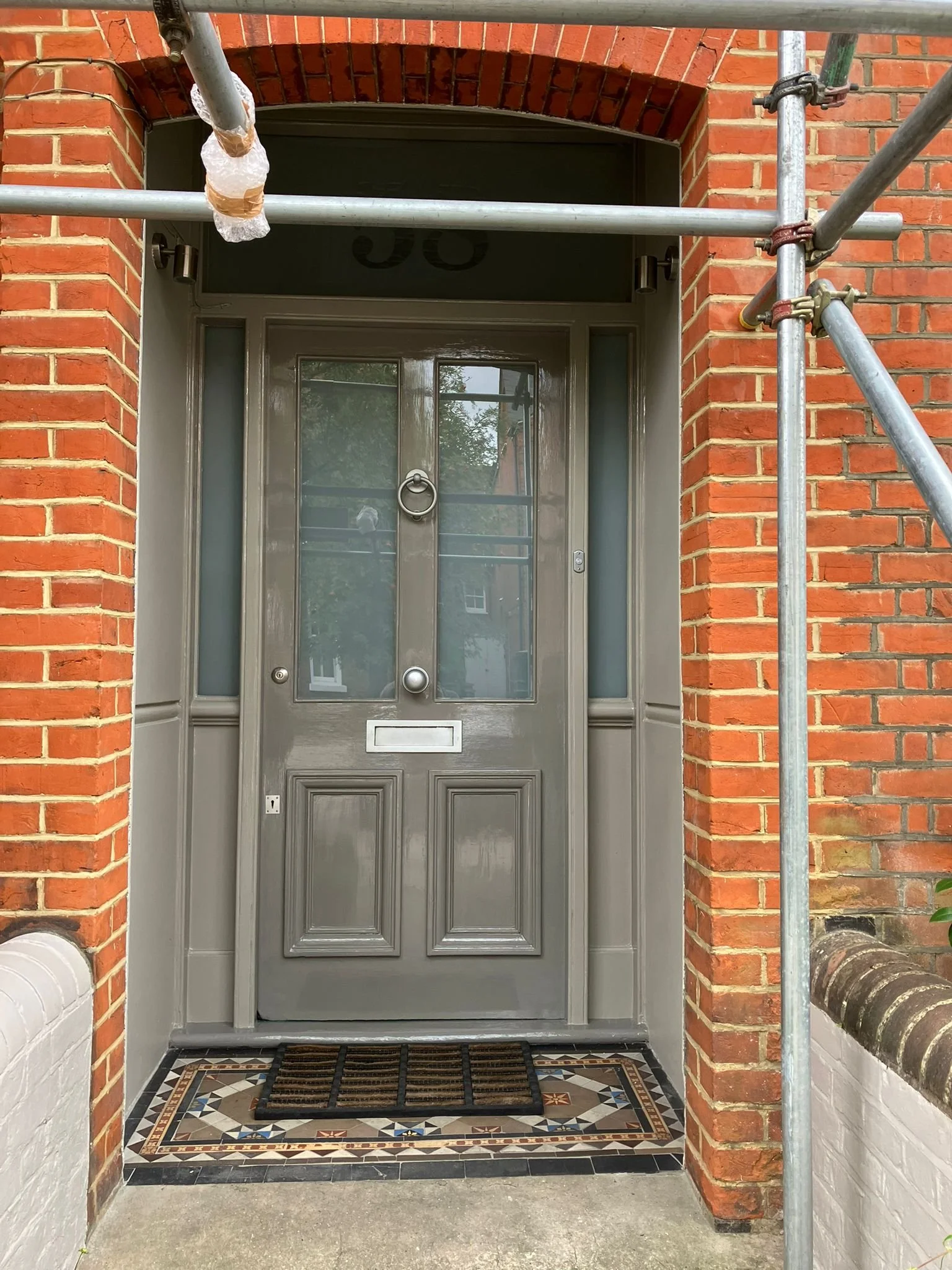 Outdoor front door of a house with red brick walls, a gray door with glass panels, a mailbox slot, and a round door knocker. There is a striped doormat and scaffolding on the right side.