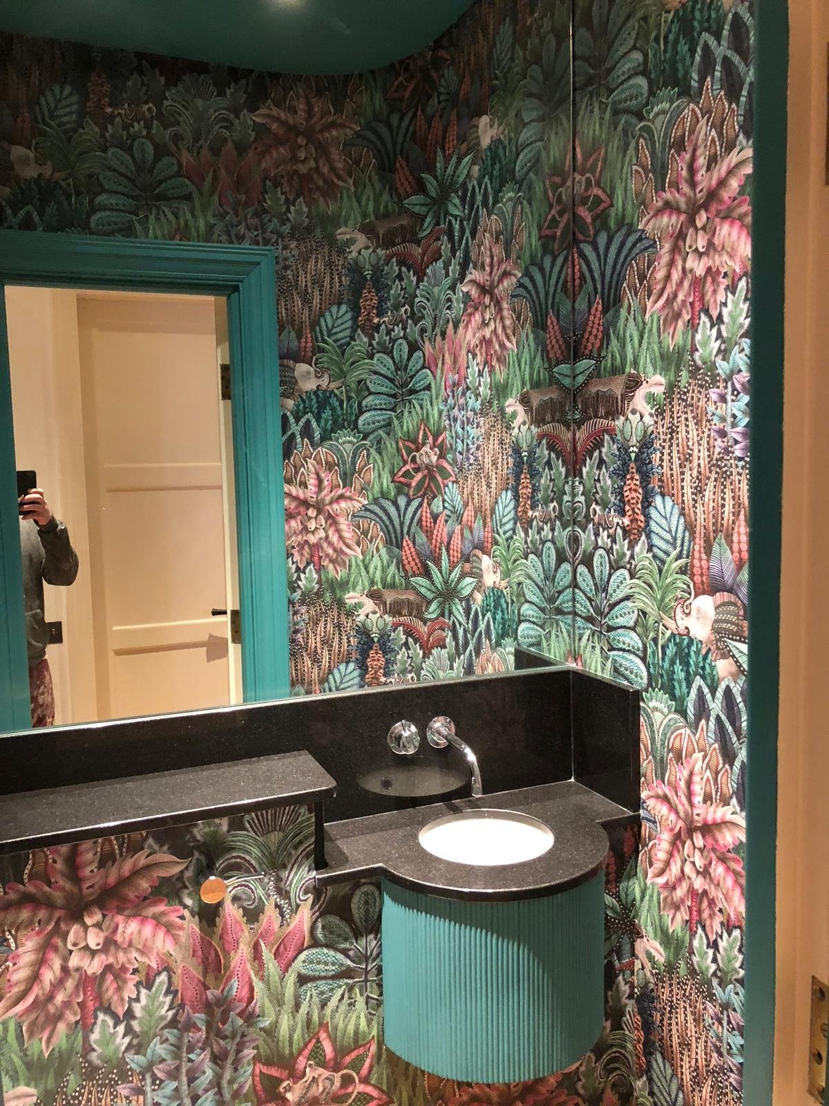 Bathroom with vibrant tropical wallpaper featuring large pink, green, and blue leaves and animals. The sink area has a black countertop with a small sink and a teal cabinet.