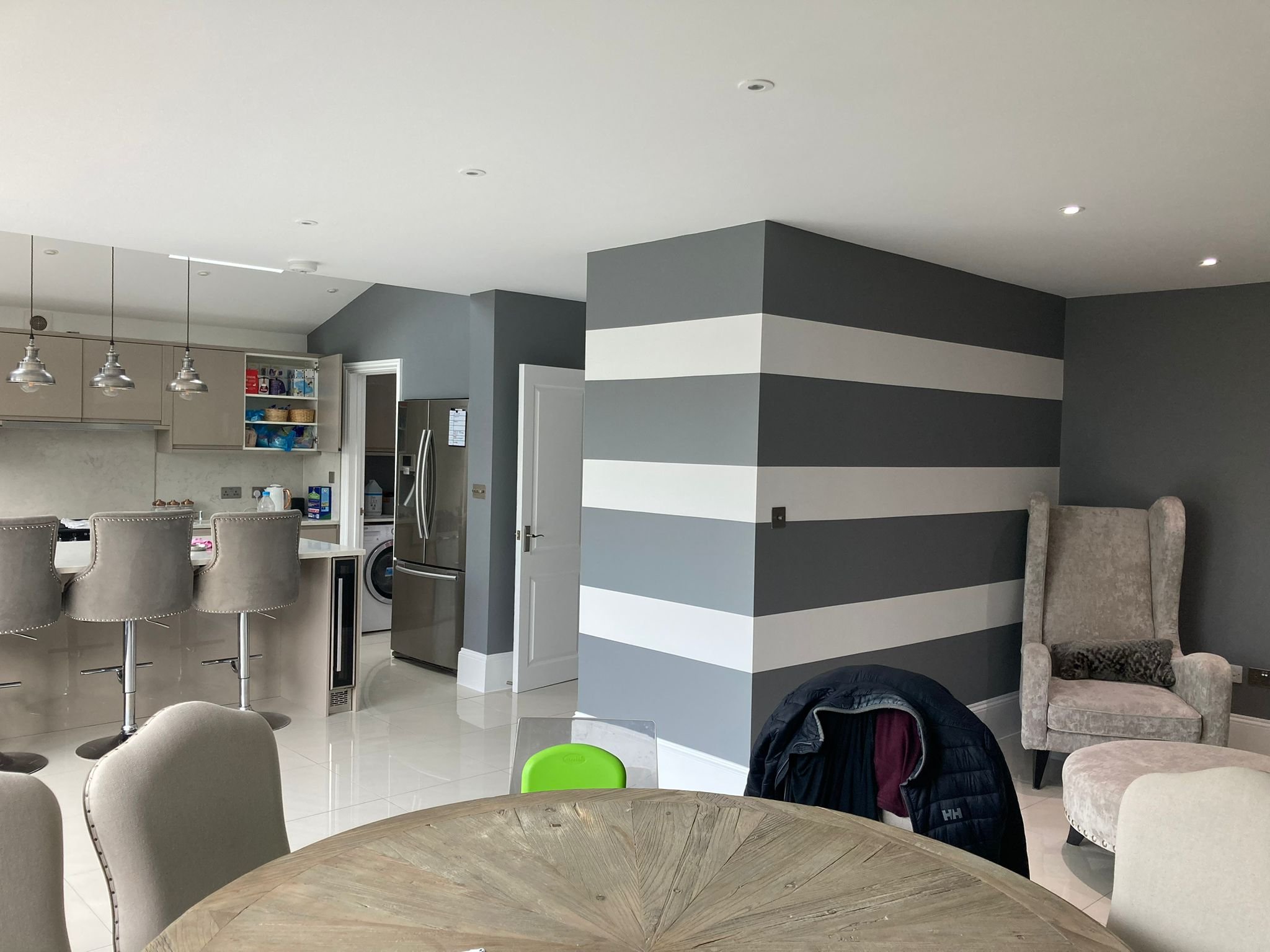 Modern open-plan kitchen and dining area with grey and white striped accent wall, beige kitchen cabinets, stainless steel appliances, kitchen island with beige barstools, and a round wooden dining table with beige chairs