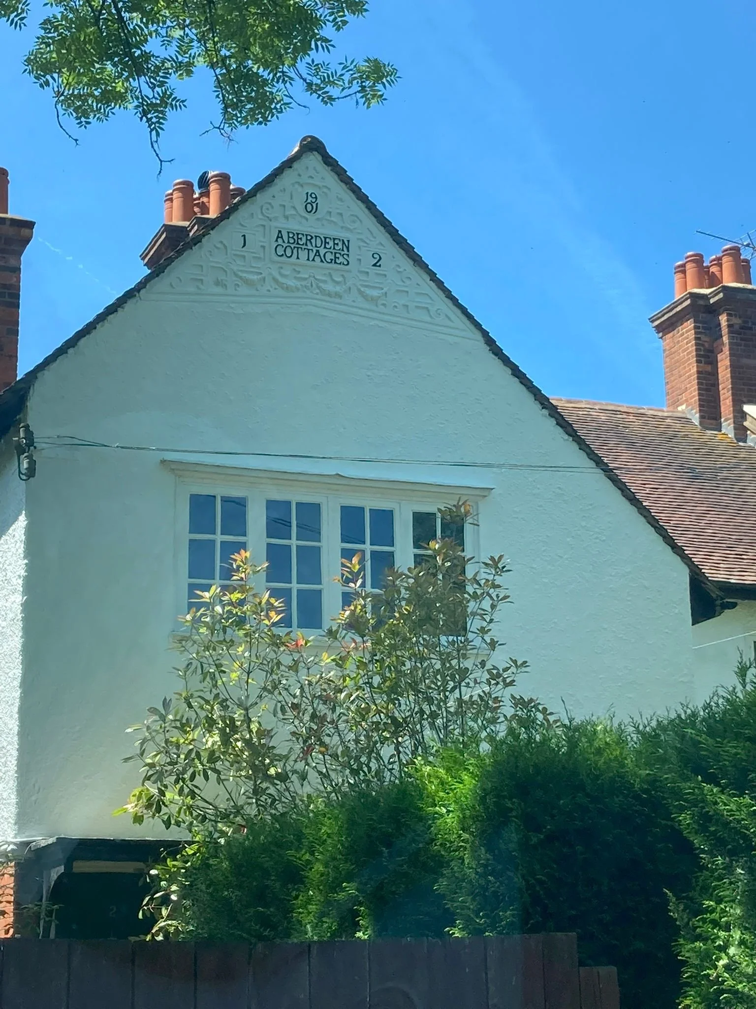 A white house with a gabled roof and a sign that reads 'Aberdeen Cottages' on the front. There are brick chimneys and a large window with multiple panes. Green bushes and trees surround the house, with a clear blue sky overhead.