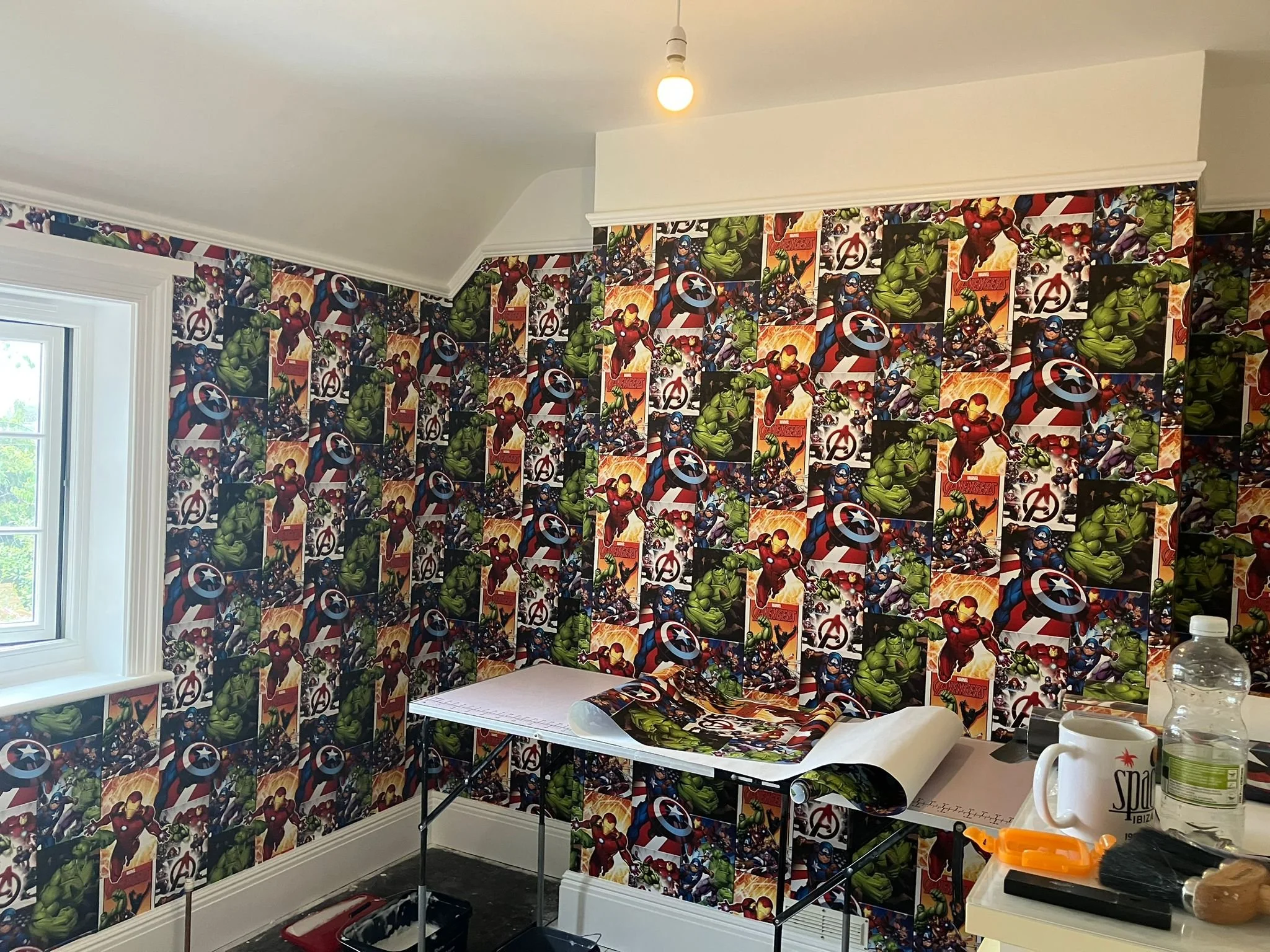 A room decorated with Marvel Avengers superhero wallpaper featuring Iron Man, Hulk, Thor, and Captain America. The room has a window, an ironing board with Marvel wallpaper paper, and a table with a coffee mug, water bottle, and miscellaneous items.