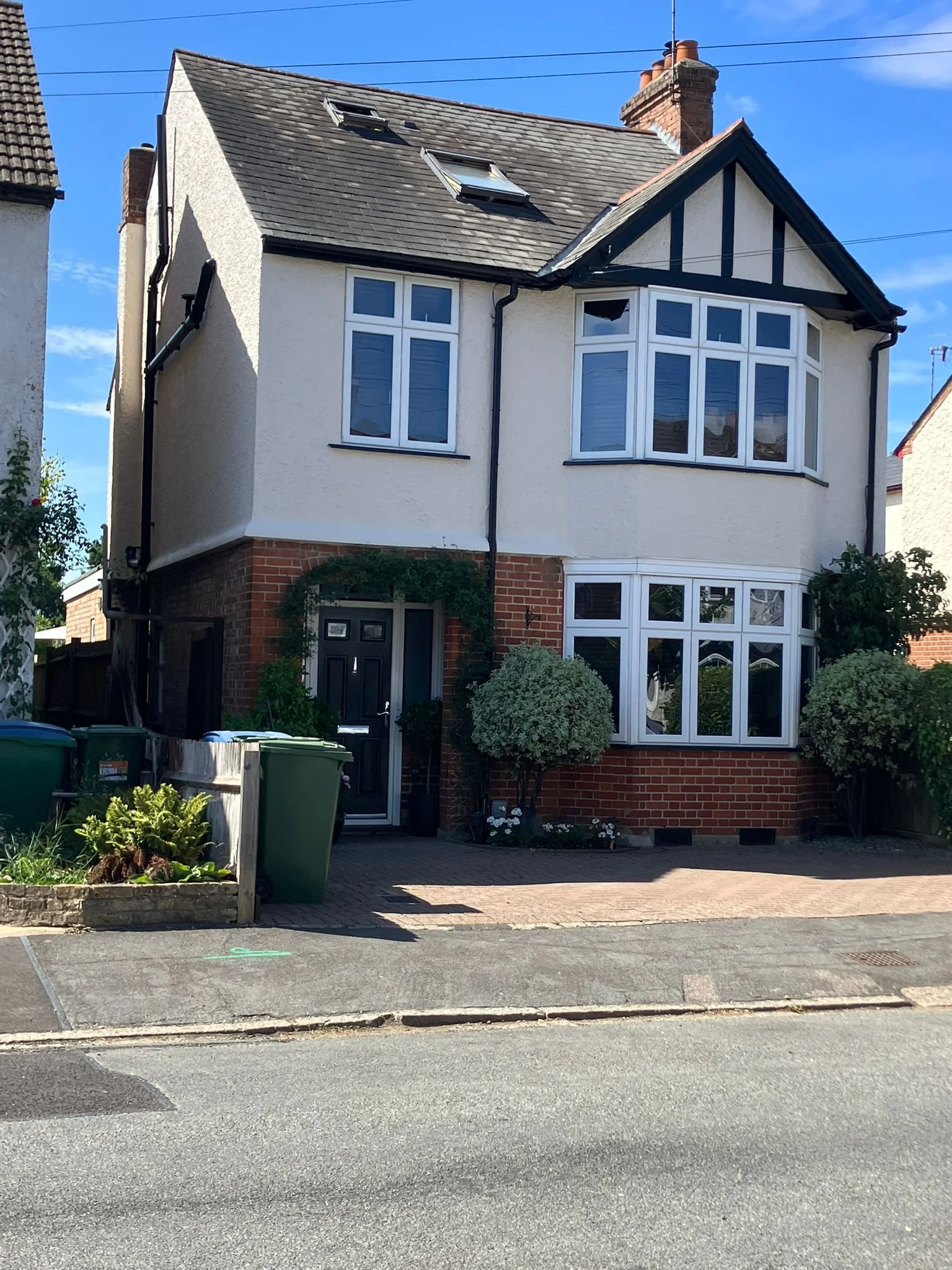 A three-story house with a brick base and cream-colored upper floors, featuring bay windows on the second and third floors, surrounded by small shrubs and trees, with a brick sidewalk in front and a clear blue sky overhead.