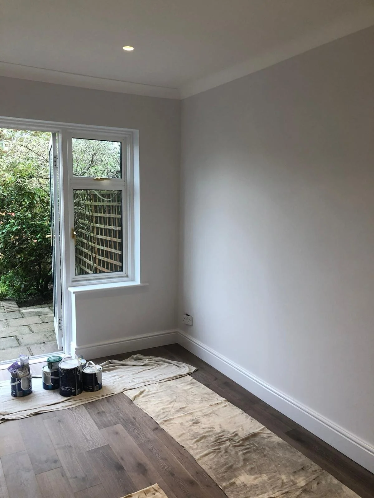 A room with new white walls, hardwood flooring, a window with a small door to a garden area, painted paint cans and fabric on the floor. The room is undergoing painting or renovation.