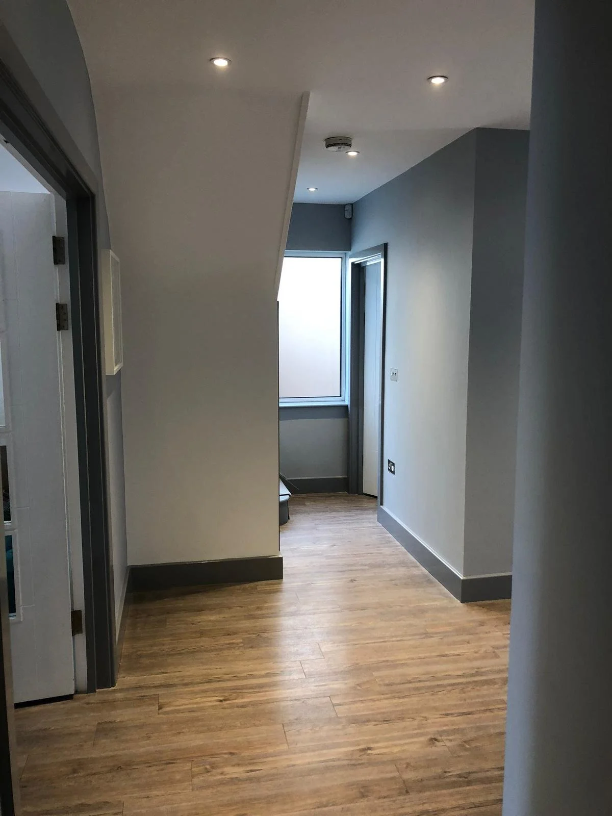 Interior corridor with wood flooring, white walls, a window at the end, and ceiling lights.