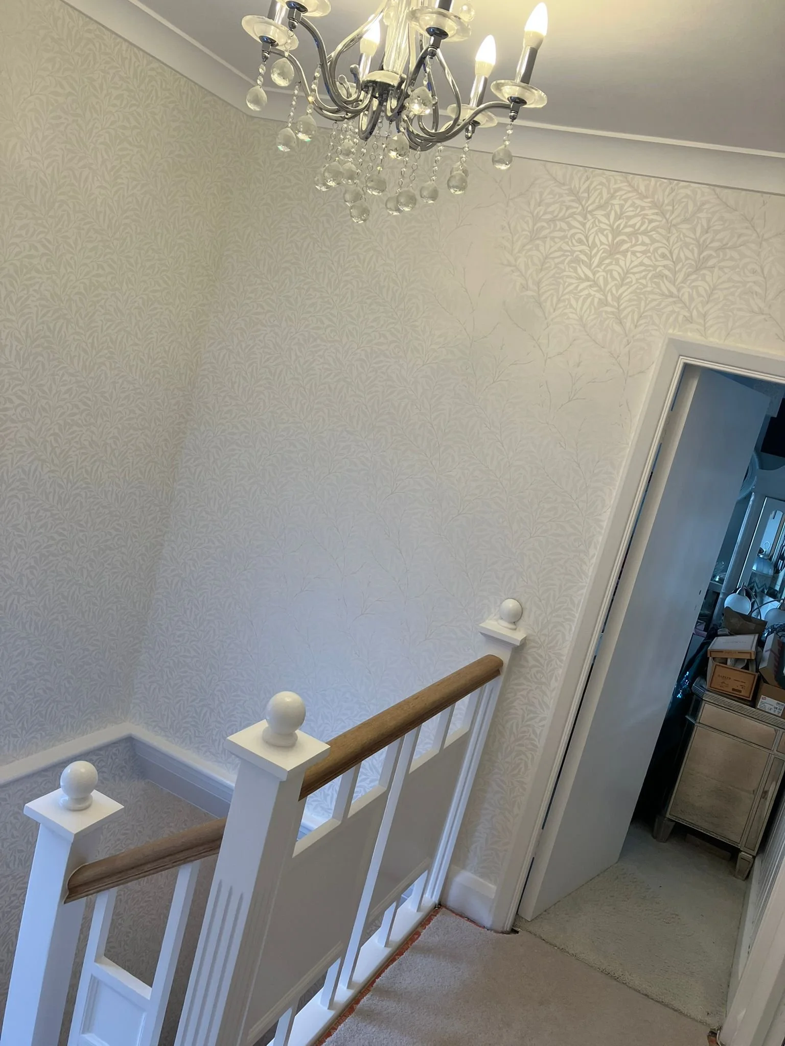 A staircase landing with a chandelier hanging from the ceiling, a wallpapered wall with a subtle leaf pattern, and a partially open door revealing storage boxes inside the closet.