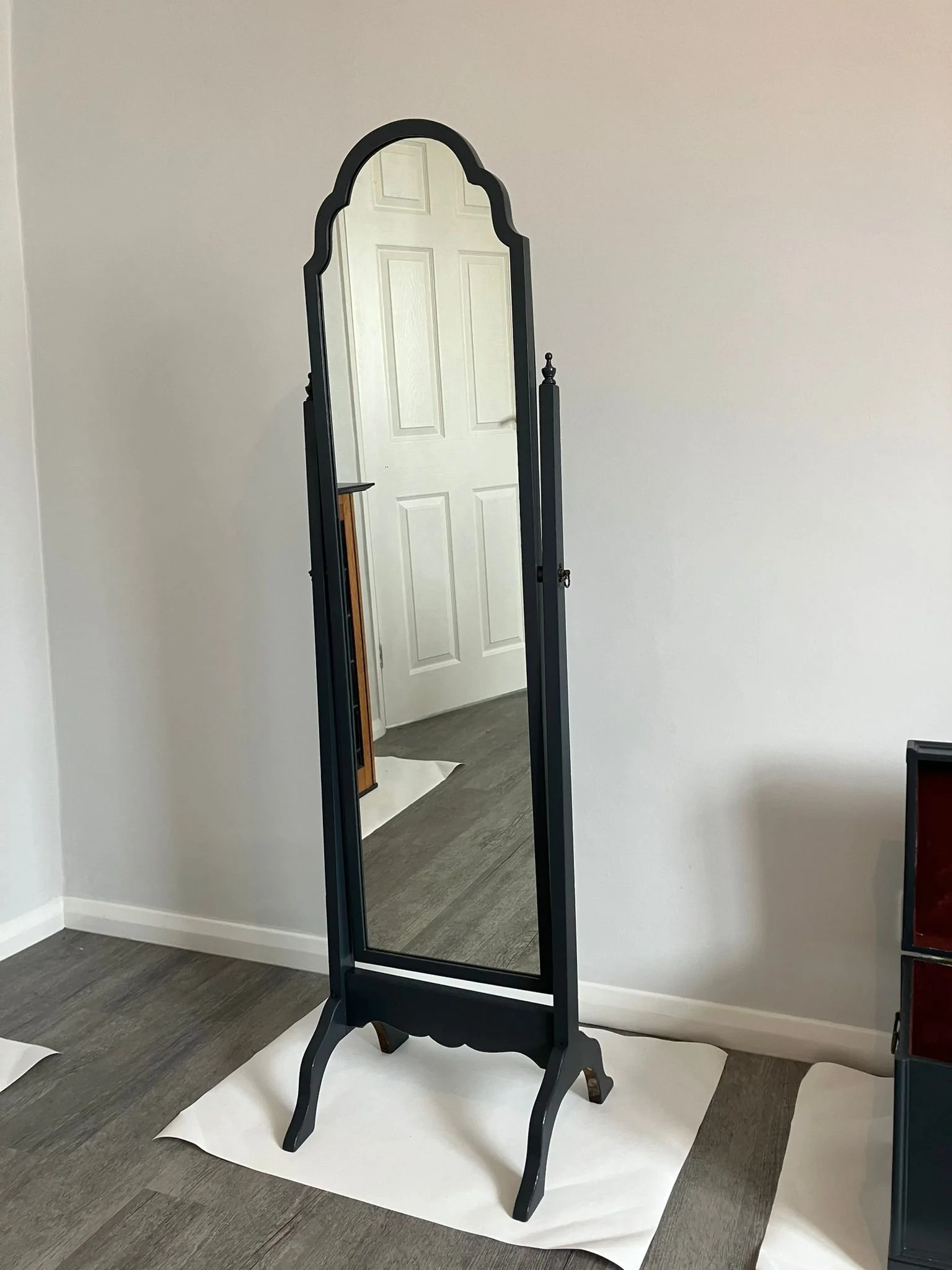 Full-length black standing mirror with an arched top, reflecting a white door and part of a room, placed on a white paper sheet on a gray wooden floor.