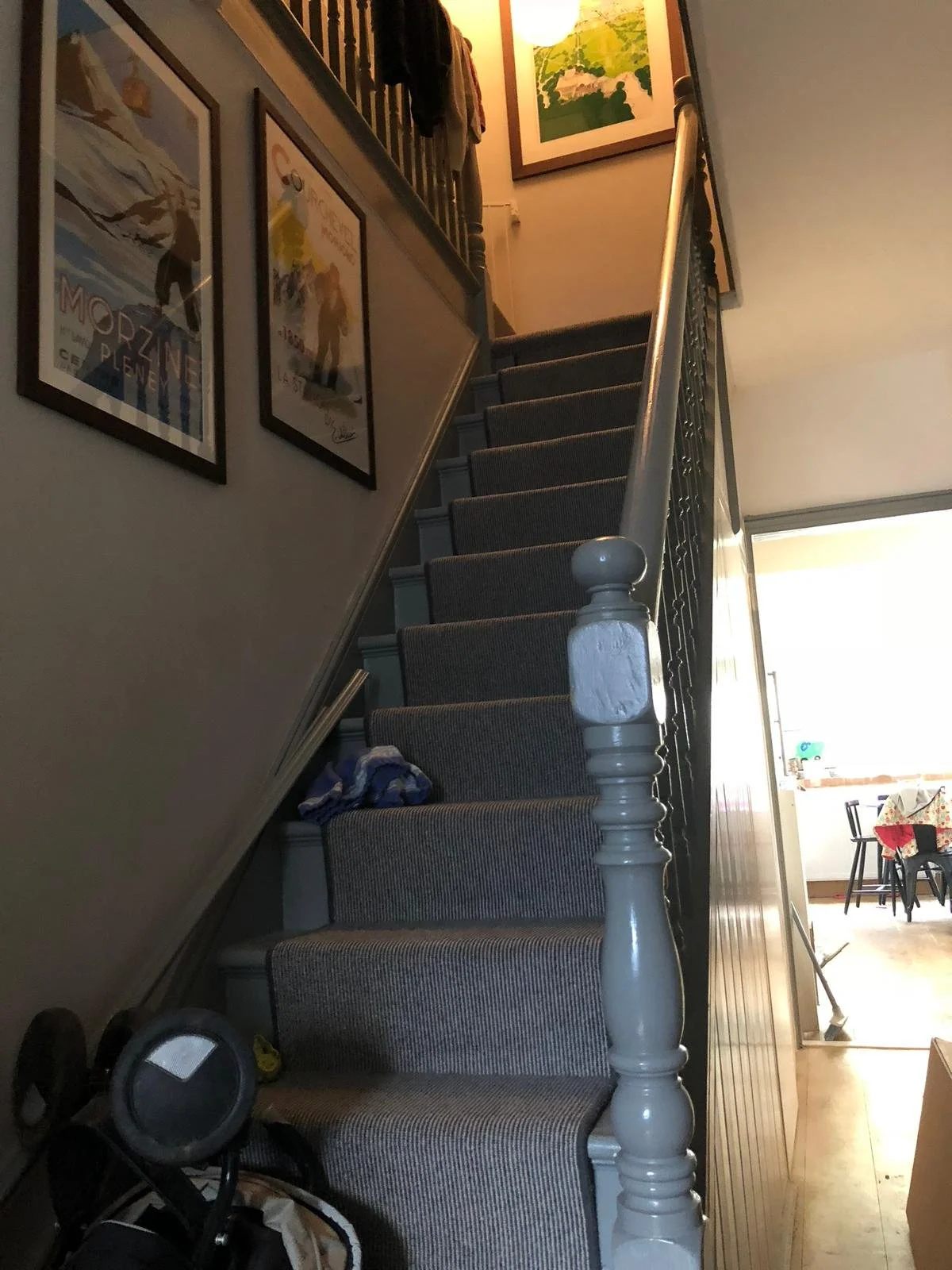 Interior view of a staircase with carpeted steps, framed posters on the wall, and a glimpse into a dining area with a table and chairs in the background.