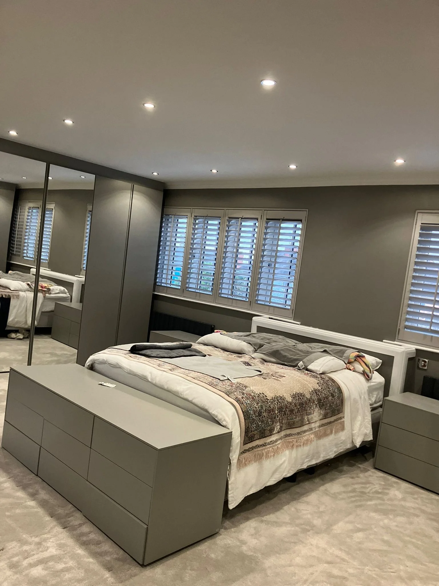 A modern bedroom with gray walls, a bed with a patterned blanket, and multiple windows with plantation shutters. There are bedside tables and a mirrored closet.