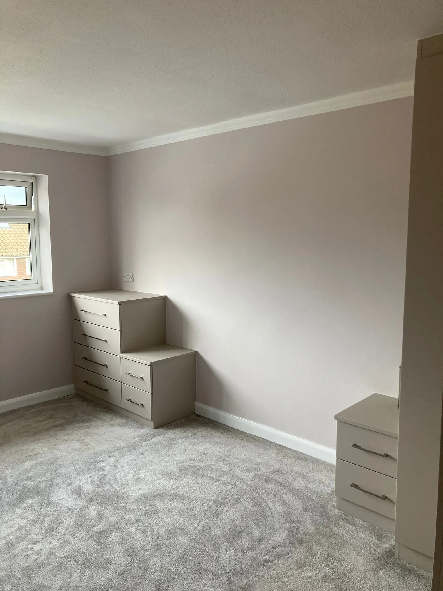 Empty bedroom with beige walls, a window, and two sets of white drawers on a carpeted floor.