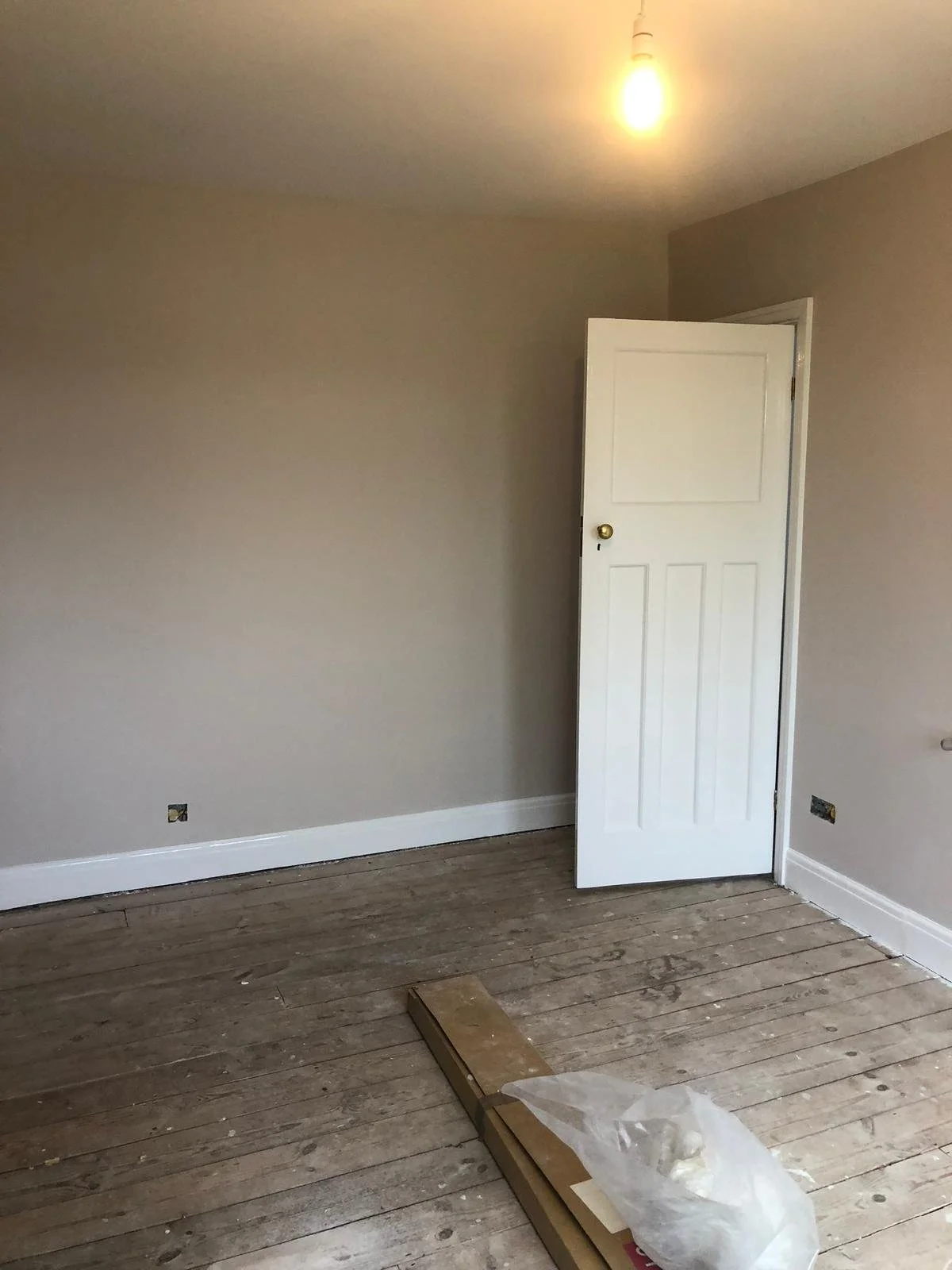 Empty room with unfinished wooden floor, gray painted walls, a white door slightly open, a lightbulb hanging from the ceiling, and some construction materials on the floor.