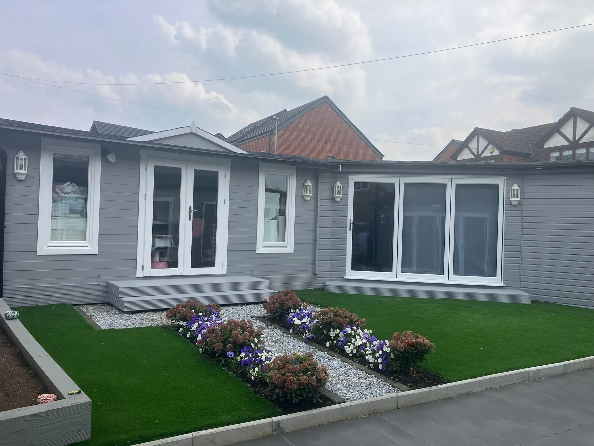 Gray house with white framed glass doors and windows, small steps leading to the door, surrounded by a lush green lawn with flower beds filled with purple, white, and brownish flowers, and a stone pathway between the flower beds, under cloudy sky.