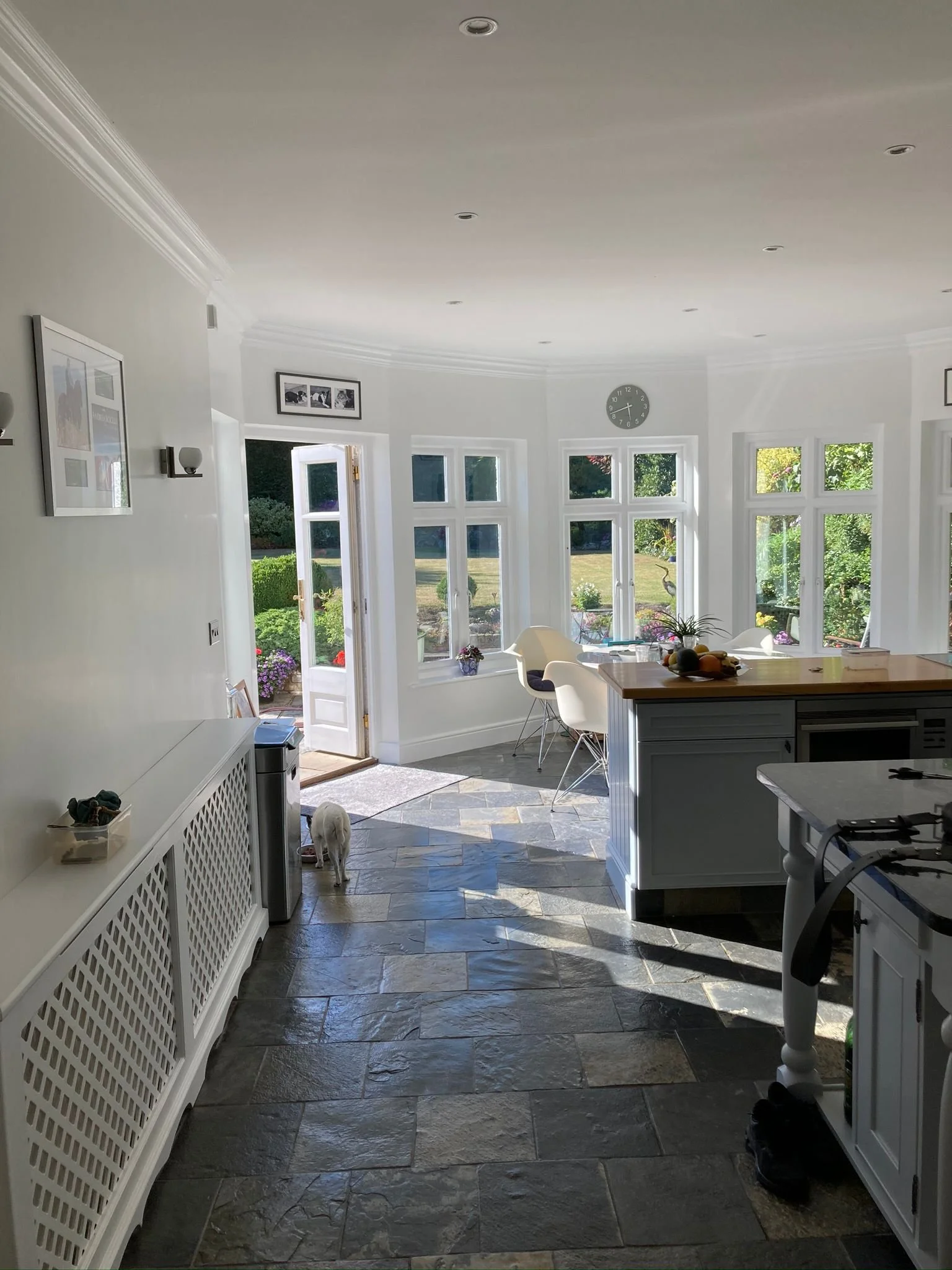 Bright kitchen with large bay windows overlooking a garden, featuring a dining table, white chairs, a kitchen island with a bowl of fruit, and a tiled floor.