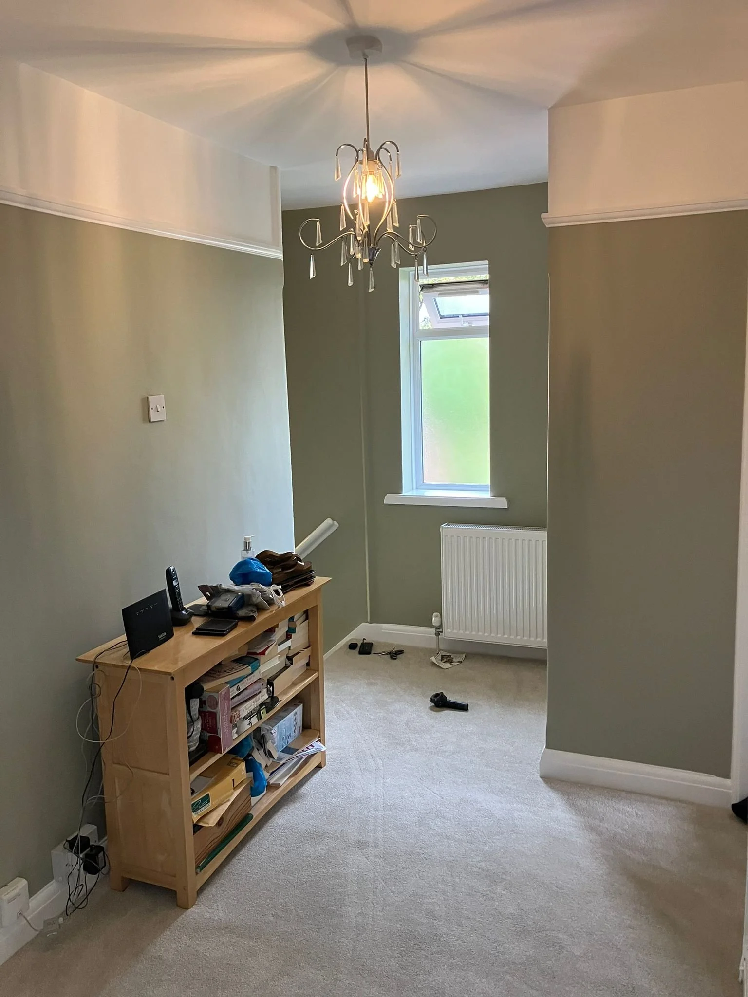 Empty room with green walls, beige carpet, a window, and a small wooden shelf with electronics and books. There is a ceiling light fixture and some tools on the floor.