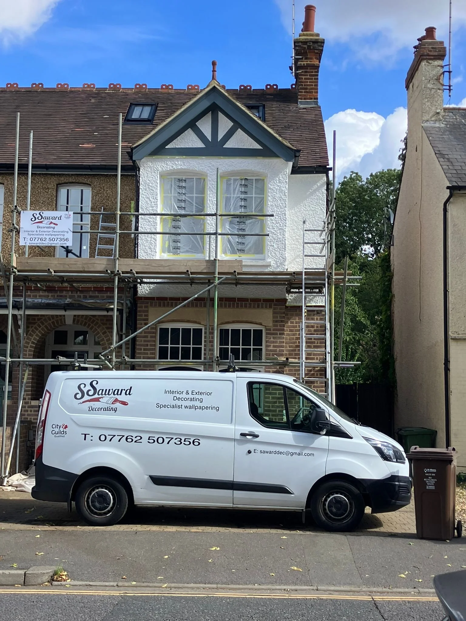 A white van with company branding parked in front of a house under renovation, with scaffolding around the house and trees in the background.
