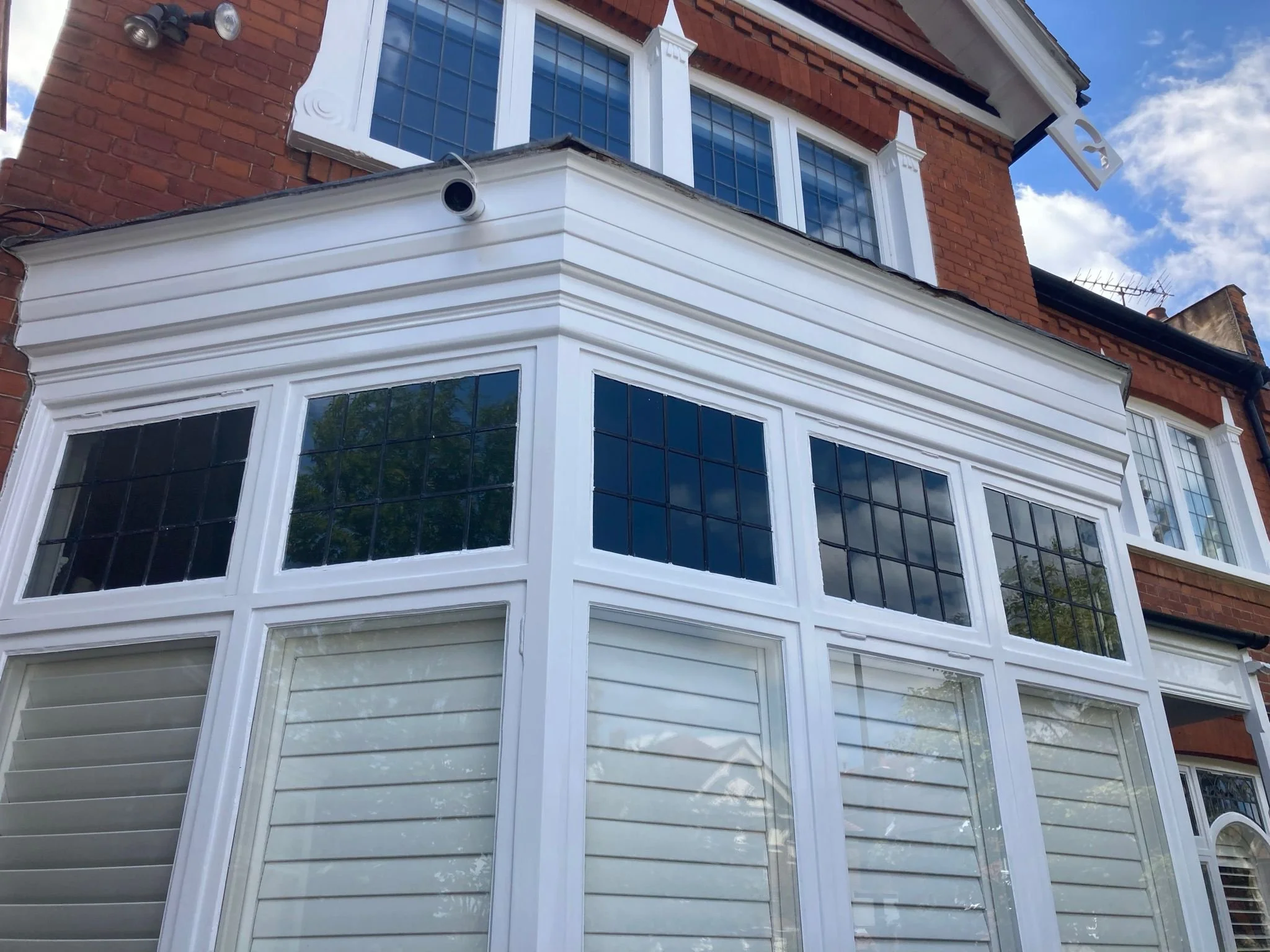 Close-up of a bay window at the front of a brick house with white trim and black window panes, with a cloudy sky above.
