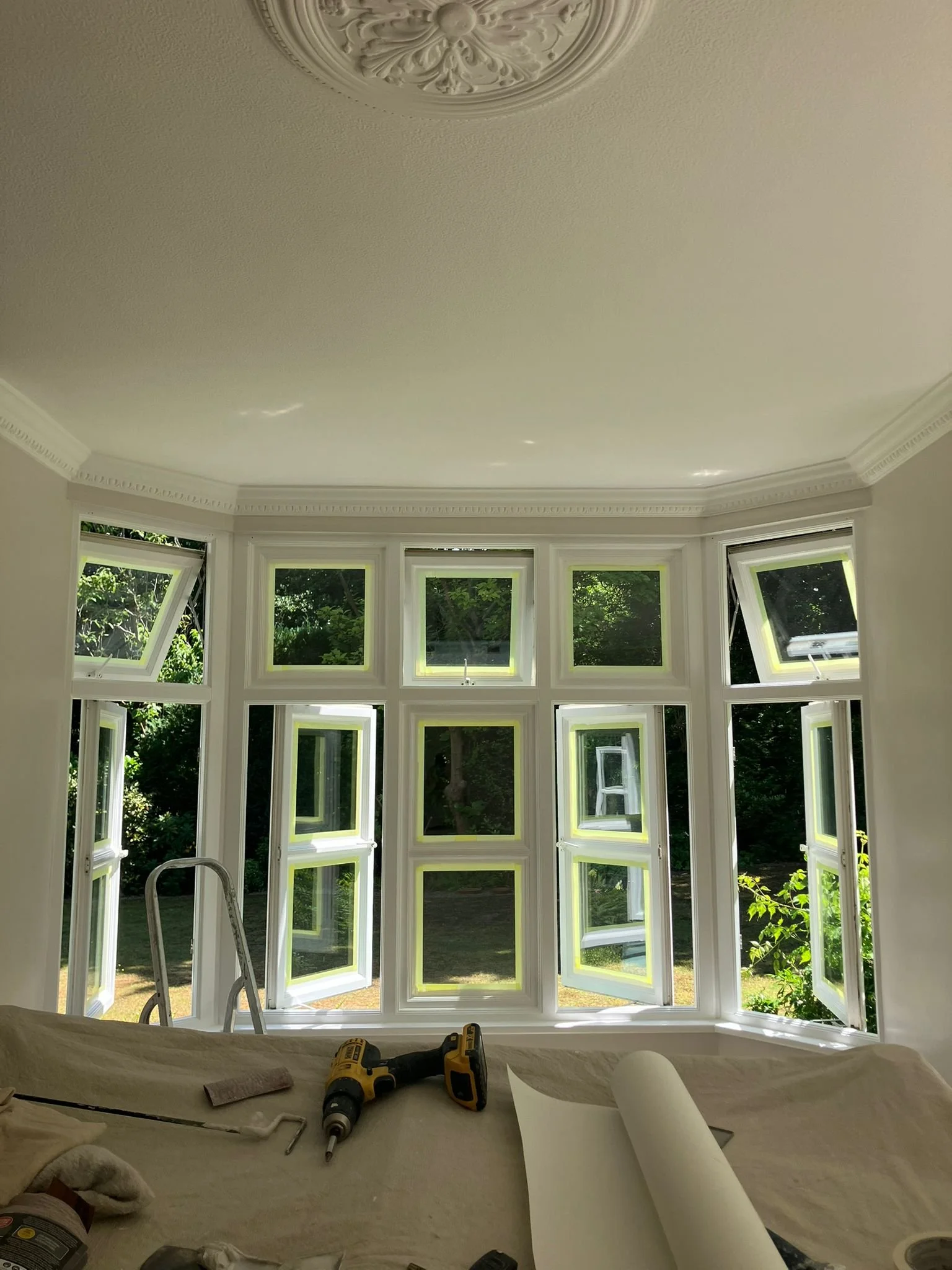 A room with a large bay window with multiple sections, some of which are open inward, revealing green trees outside. Construction tools and materials are on a sheet-covered surface in the foreground, indicating ongoing window installation.
