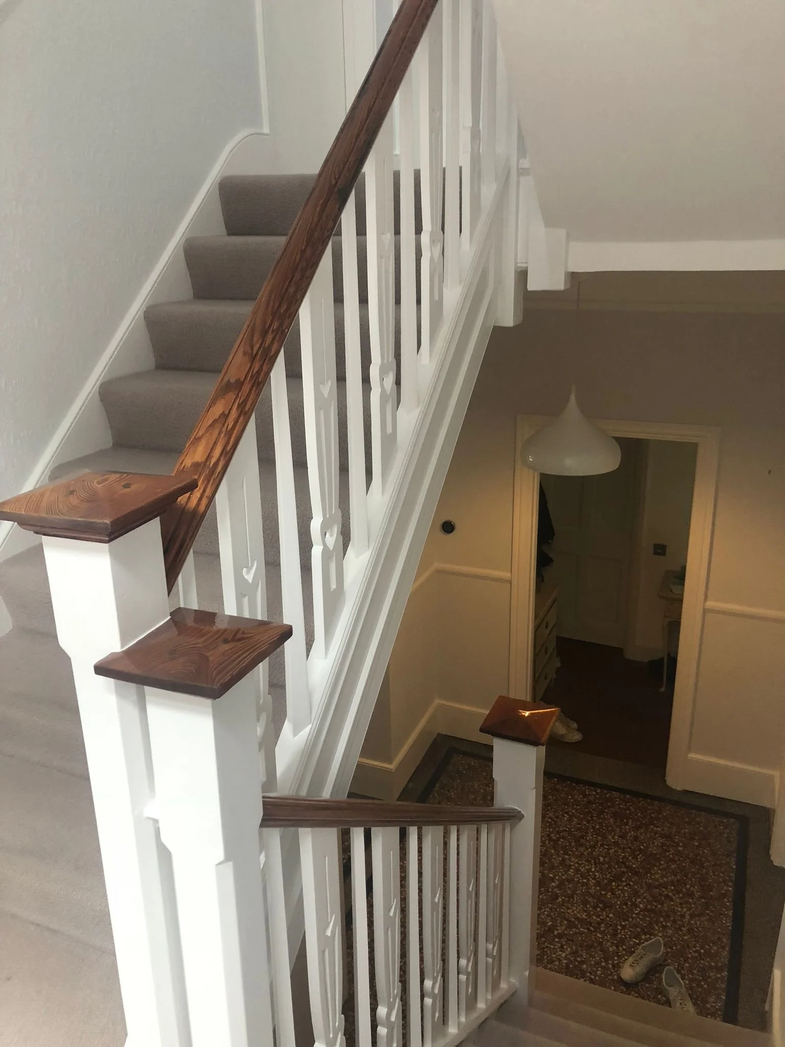 Interior view of a staircase with white railings and wooden handrails and newel posts. Carpeted stairs lead down to a lower floor with a patterned carpet and a pair of shoes. A hanging light fixture is visible on the ceiling below.