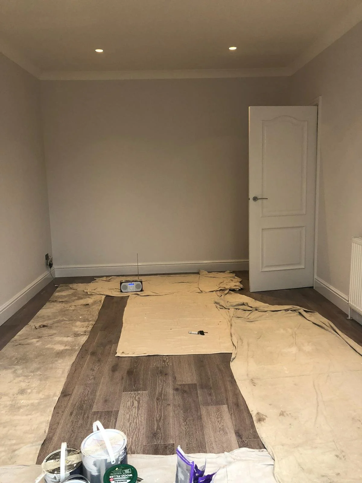 Room with partially installed wooden flooring and beige coverings on the floor. A radio and some tools are present on the floor, with a bucket and cans at the front. Walls are painted in light color, with integrated ceiling lights visible.