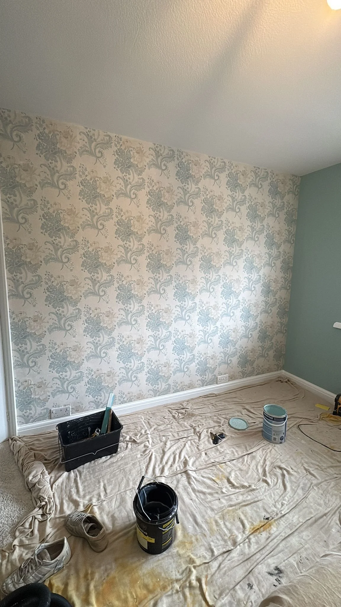 Room under renovation with painting supplies, shoes, and roll of wallpaper on the floor, decorated with floral patterned wallpaper on one wall.