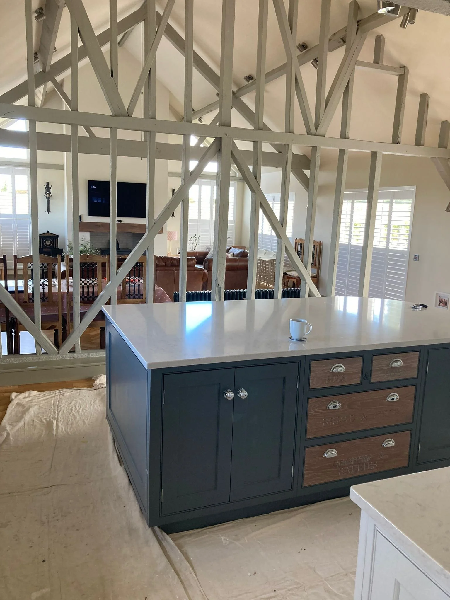 Interior view of a house with kitchen and living room, showing an unfinished wall with exposed wooden framing, a countertop with a white mug, and furniture including a dining table, chairs, and a sofa set in the background.