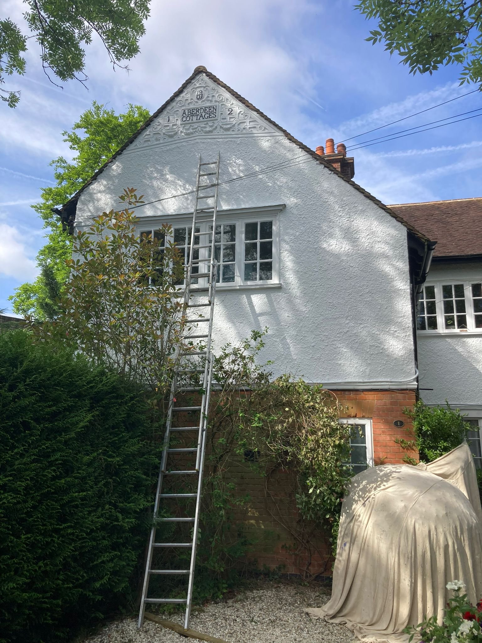 White house with cloudy sky, bush, ladder, and covered object outside.