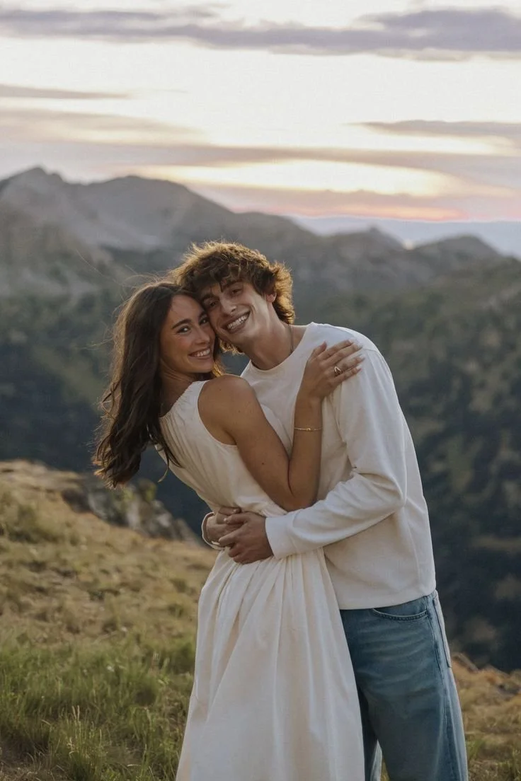 Un couple souriant en étreinte dans un paysage montagneux au coucher de soleil.