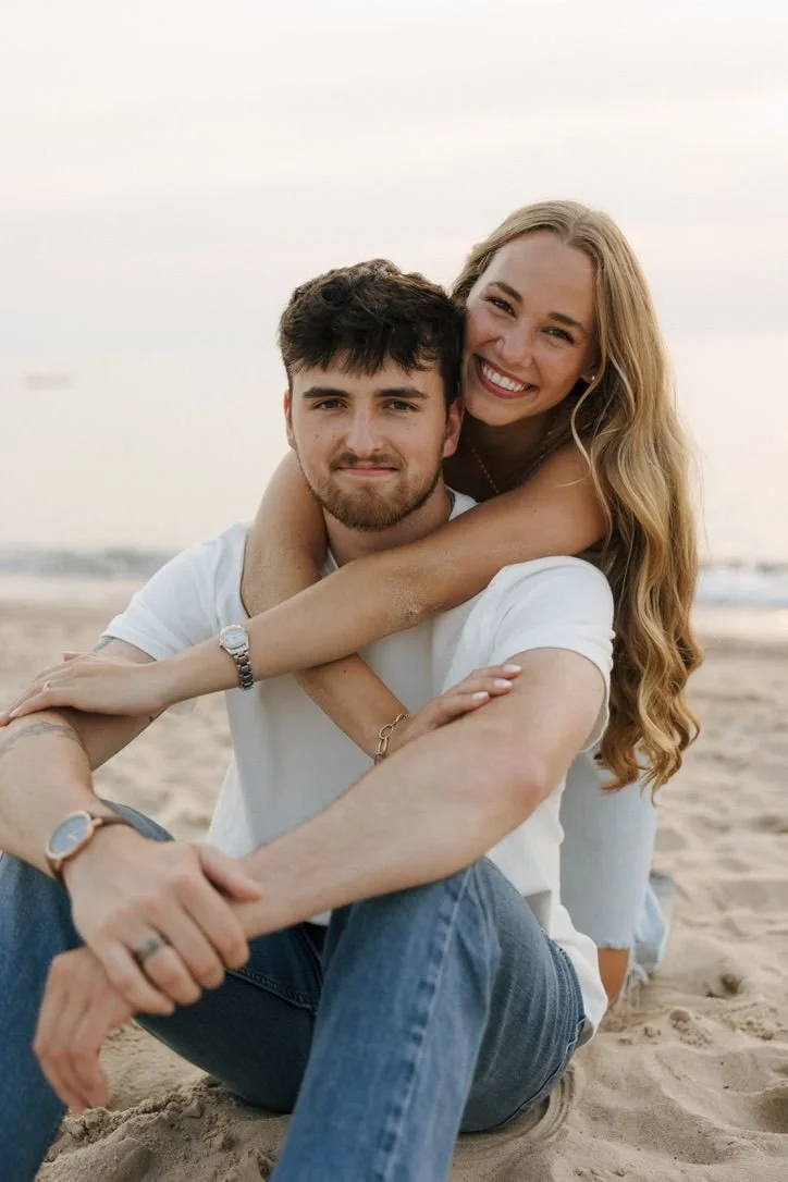 Un couple souriant assis sur la plage, avec la femme qui embrasse le homme par derrière, dans un décor de plage avec la mer en arrière-plan.