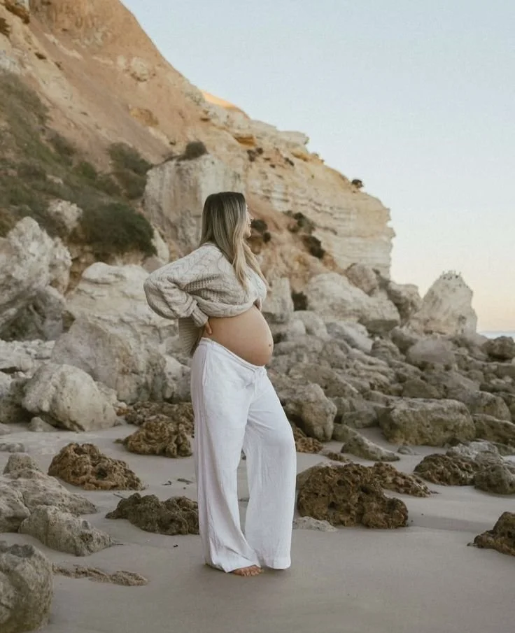 Une femme enceinte debout sur la plage, regardant vers la droite, avec un paysage rocheux en arrière-plan.