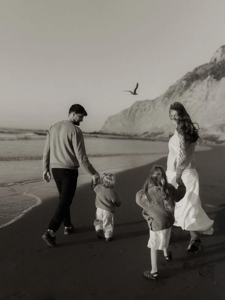 Une famille marche sur la plage au coucher du soleil, avec des falaises en arrière-plan, en noir et blanc.