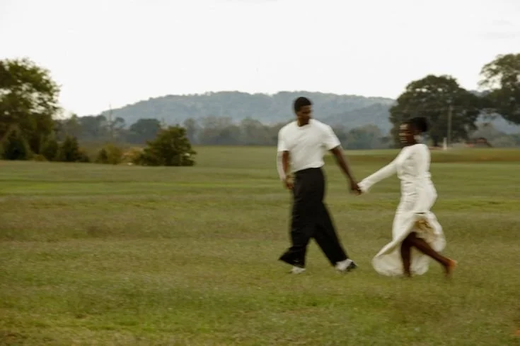Un couple marche dans un champ vert, se tenant par la main, avec des arbres et des collines à l'horizon.