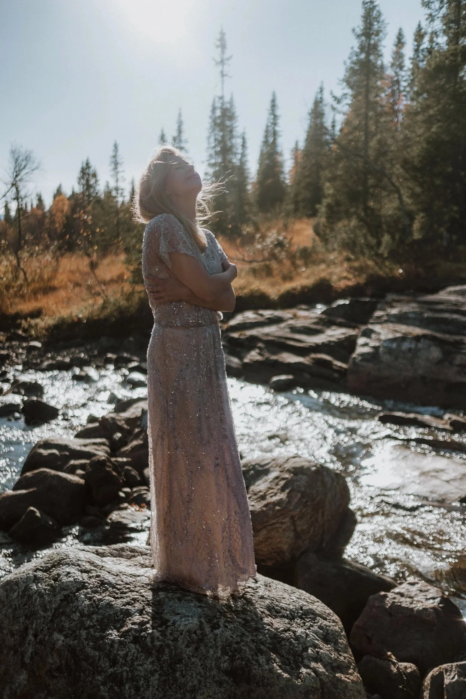 Olga in a sparkly pink dress standing on a large rock by a river with trees in the background, enjoying sunlight.