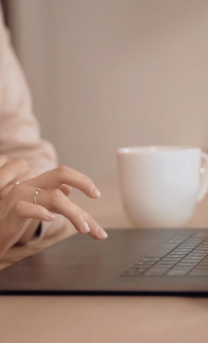 A woman's hand typing on a laptop with a white mug in the background.