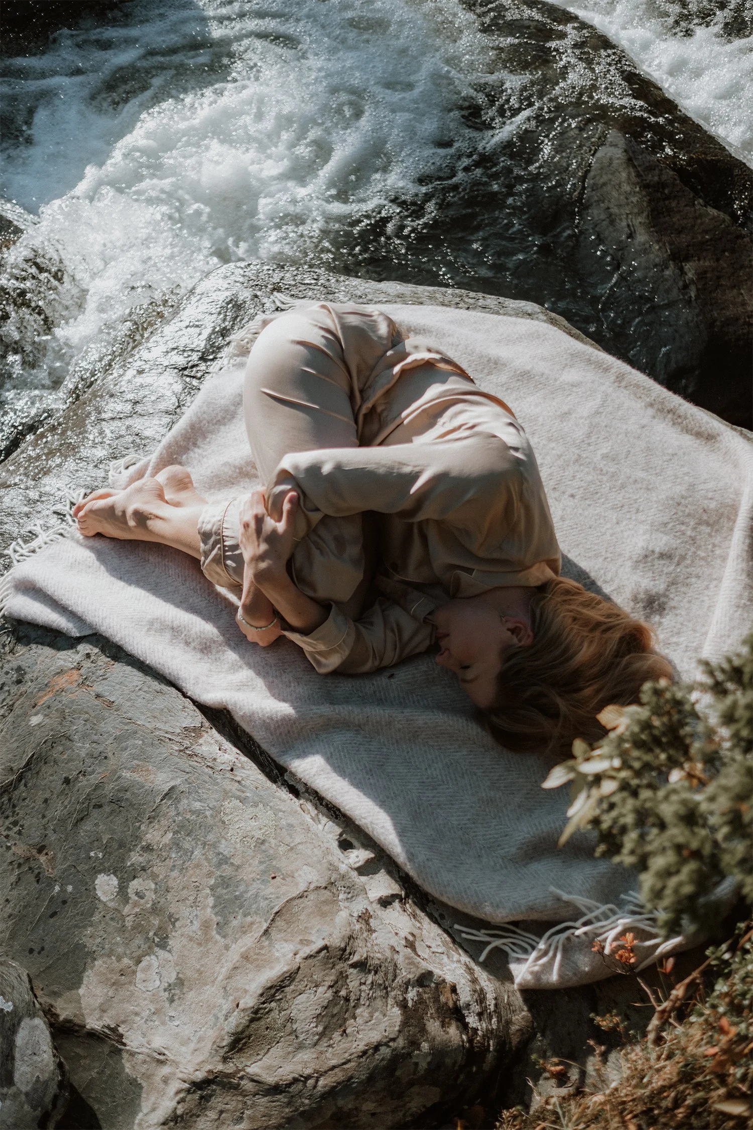 Olga curled up on a giant rock, hugging her knees with her eyes closed, resting near a beautiful, blue, flowing river.