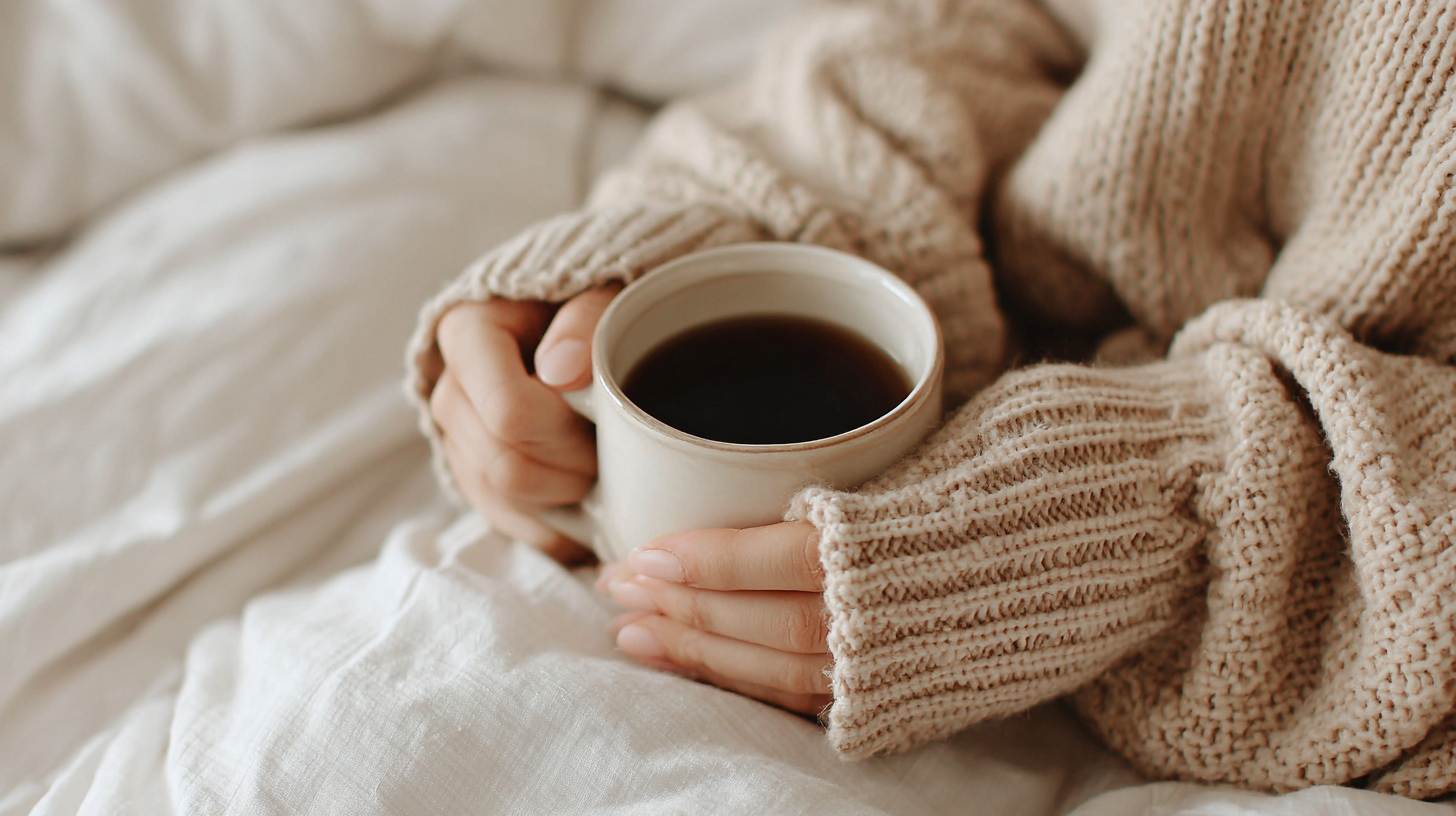 Person in cozy beige sweater holding a white mug of black coffee while sitting on a bed with white linens.