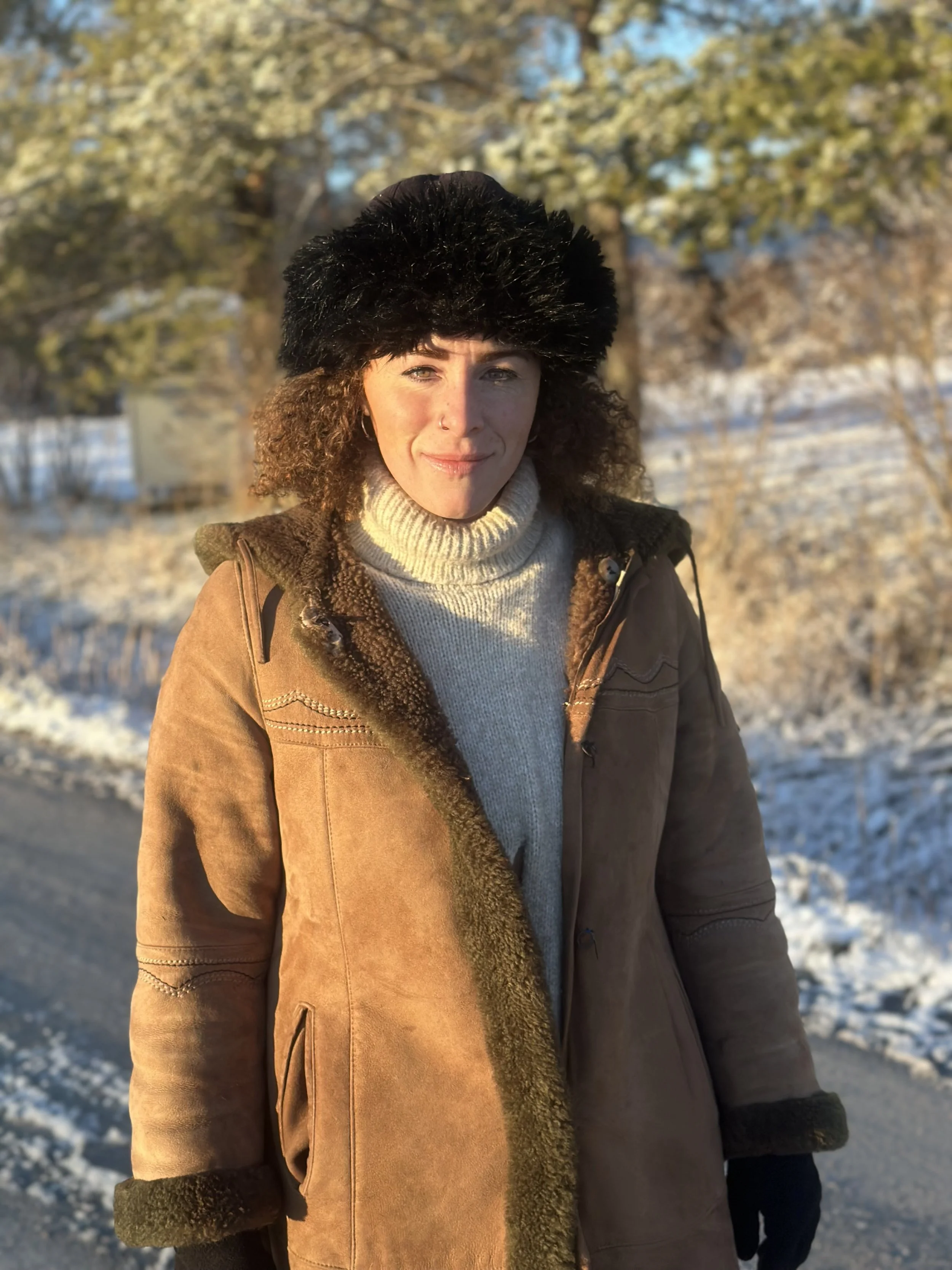 Ciara, a woman with brown curly hair standing outdoors in winter, wearing a black fur hat, and a brown coat, with snow-covered ground and trees in the background.