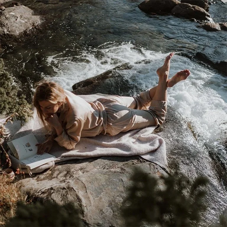 Olga lying on her stomach on a blanket by a river, reading a book, with her legs bent and feet in the air.