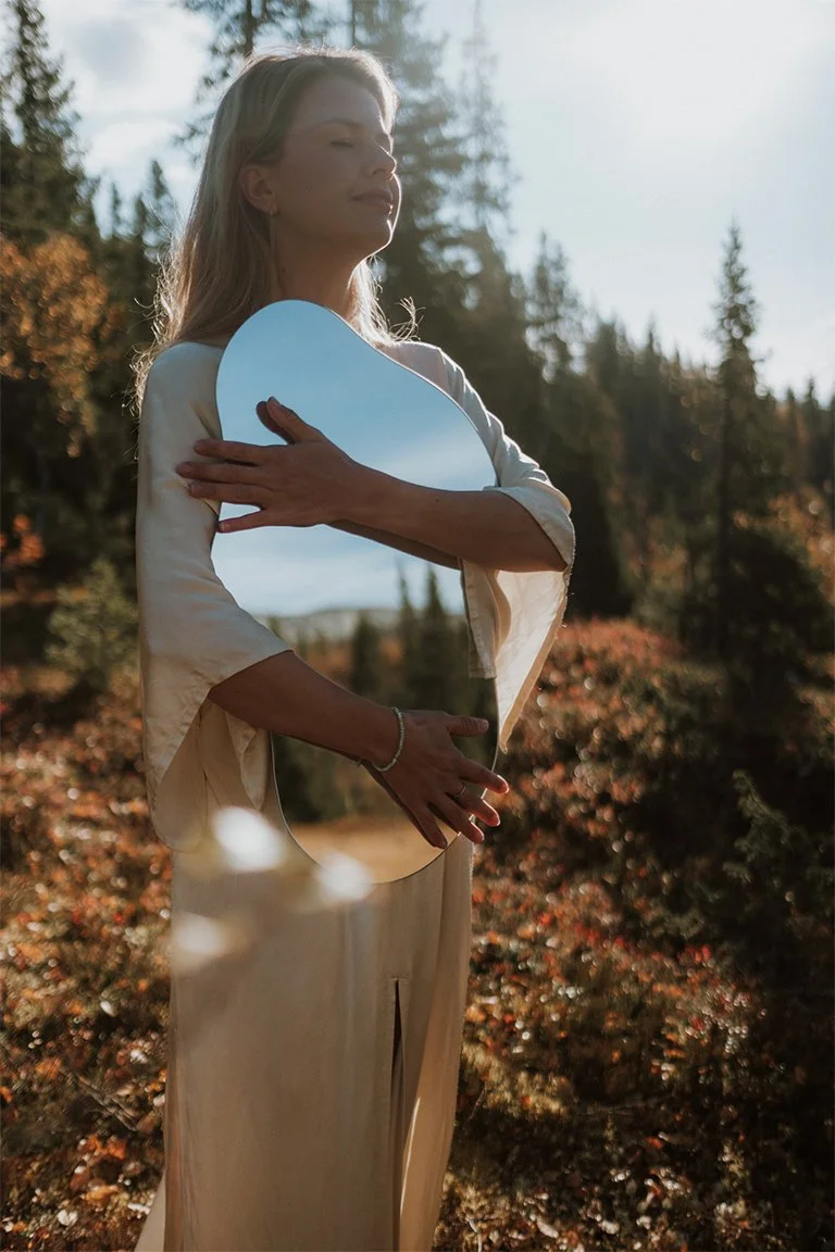 A peaceful Olga with her eyes closed, holding a rounded organic mirror against her torso, with trees autumn foliage, and sky  reflected in the mirror.