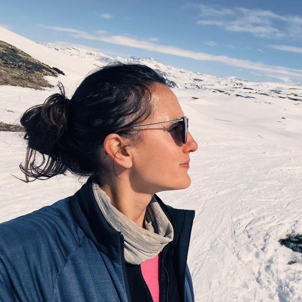 Lena, a woman with brown hair wearing sunglasses and a blue jacket standing in a snowy mountain landscape with clear blue skies.