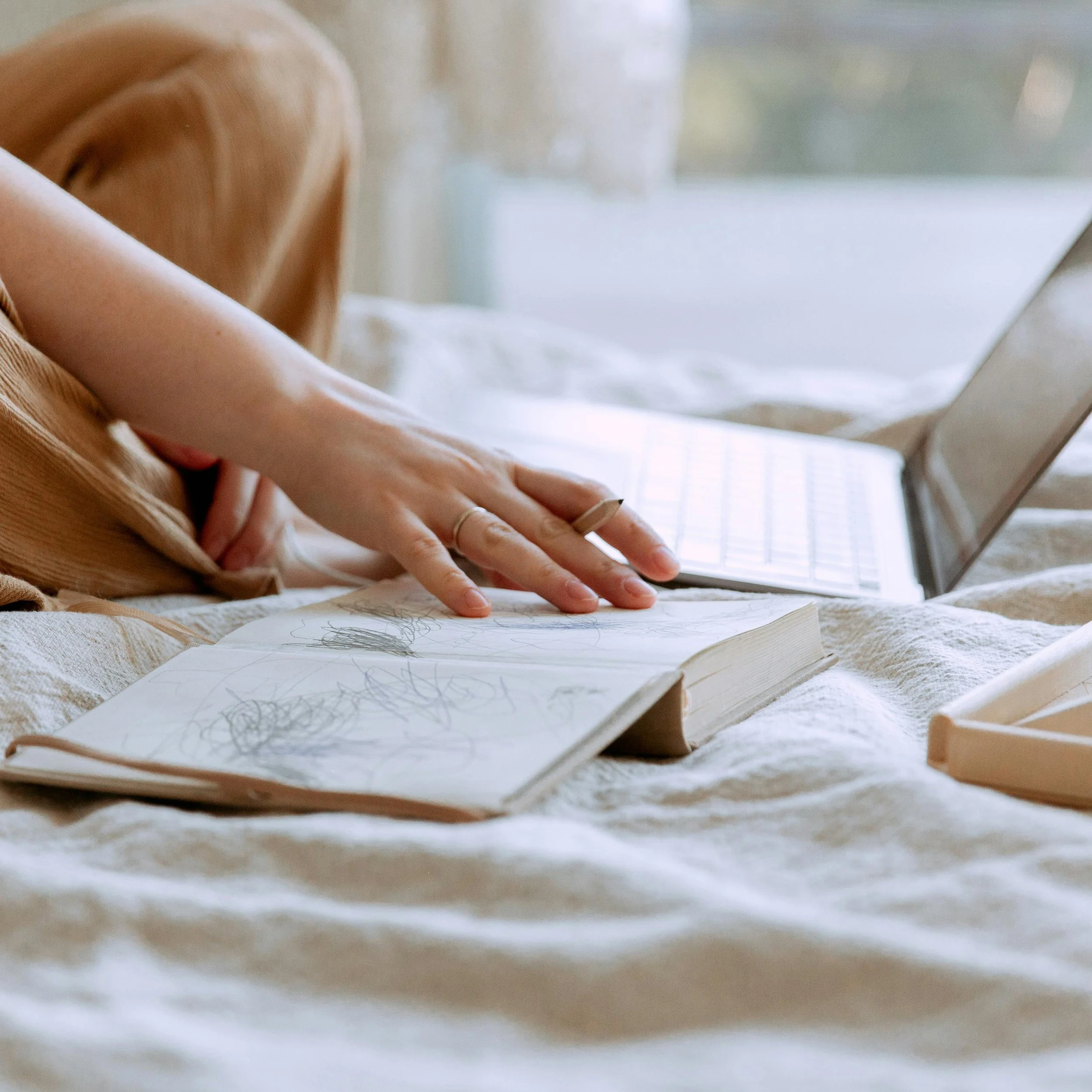 A person sitting criss-cross on a bed, writing or drawing in a notebook, while a laptop is open nearby. The person is wearing rings and beige clothing.