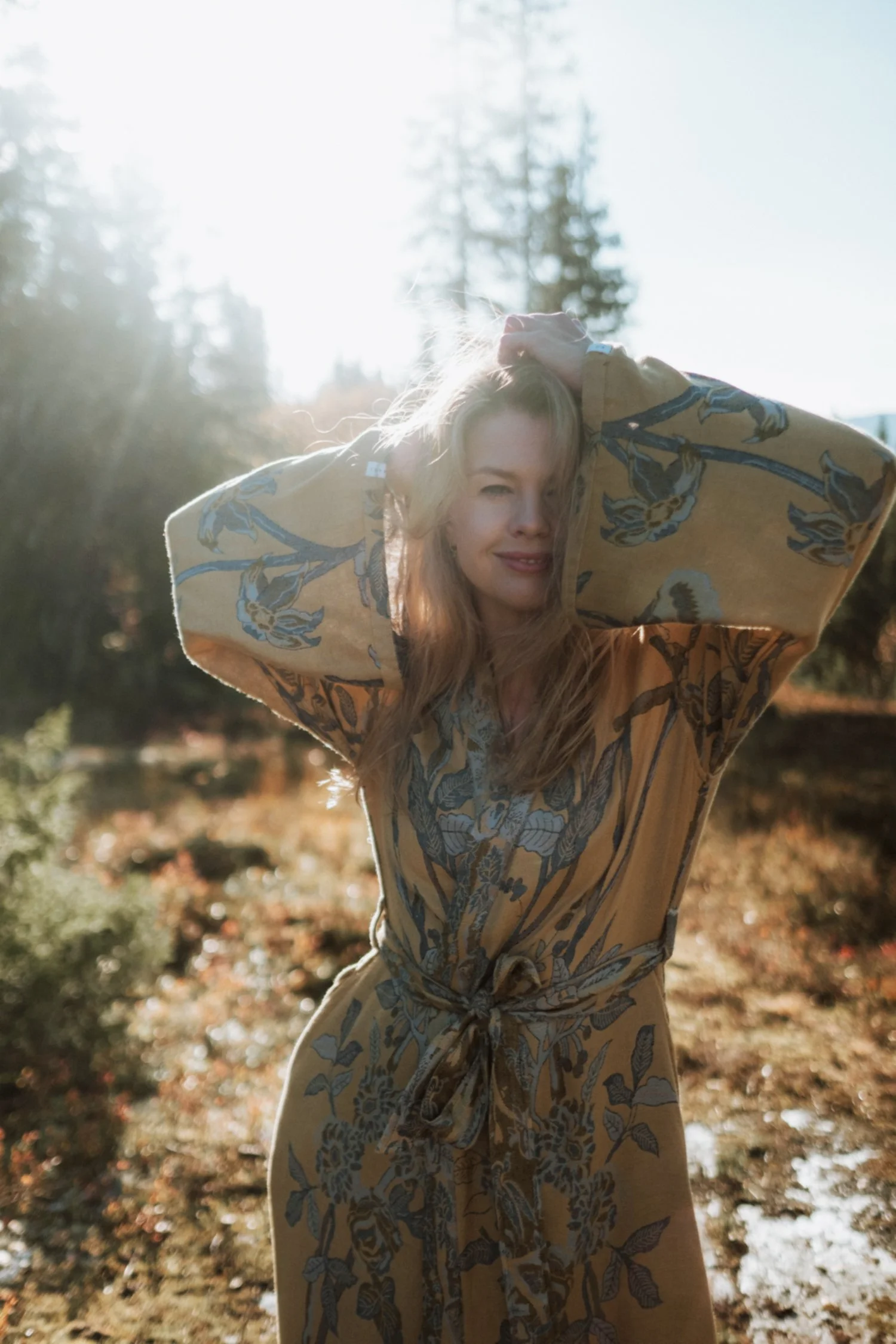 Olga outdoors in sunlight, wearing a floral patterned dress, with her hands in her hair, standing amongst trees.