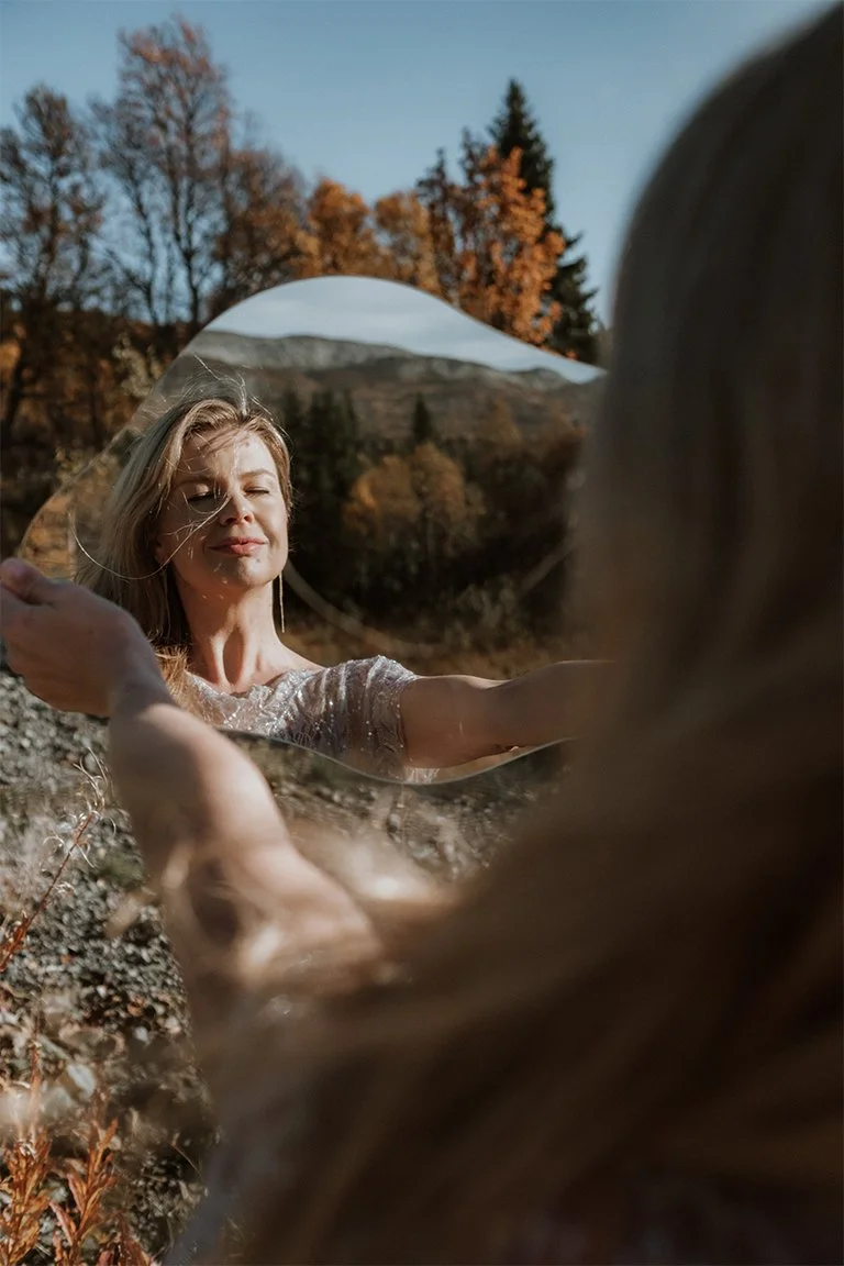 Olga holding a mirror in front of her and the reflection has herself, autumn trees and mountains.