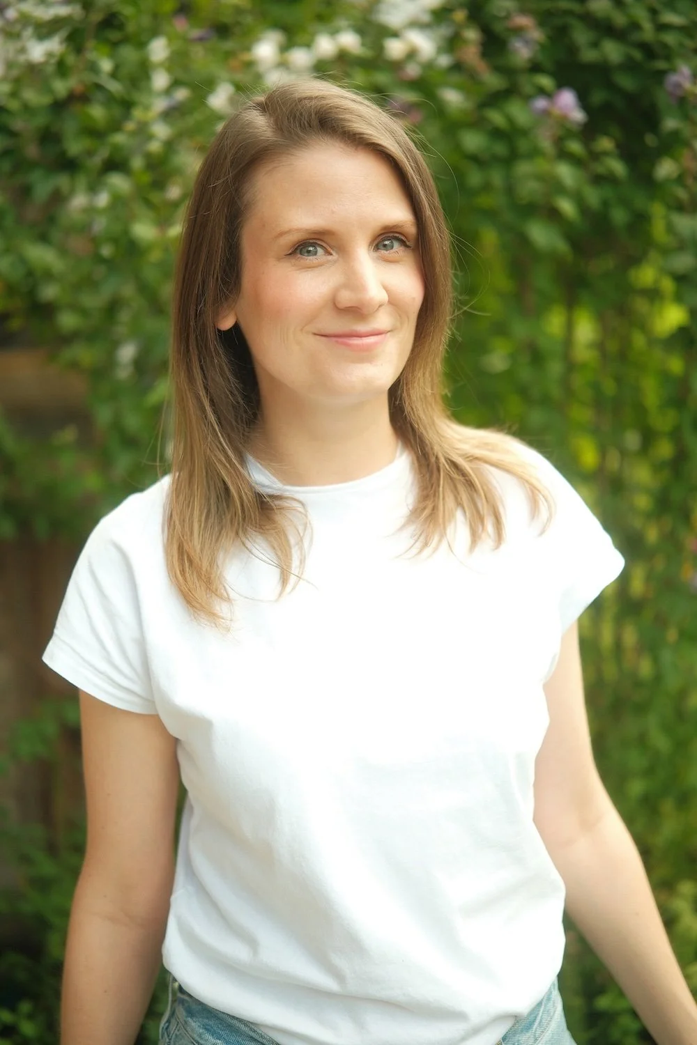 Doula mit braunen Haaren in einem weißen T-Shirt vor einer grünen Gartenkulisse. Doula Begleitung Berlin