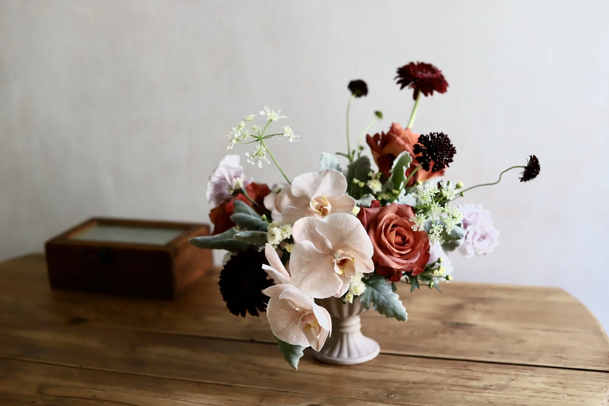 A floral arrangement in a white pedestal vase placed on a wooden table, with beige and brown flowers and greenery.