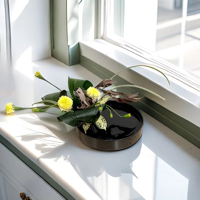 Decorative flower arrangement with yellow chrysanthemums and mixed foliage in a black round vase on a white windowsill with sunlight and shadows.