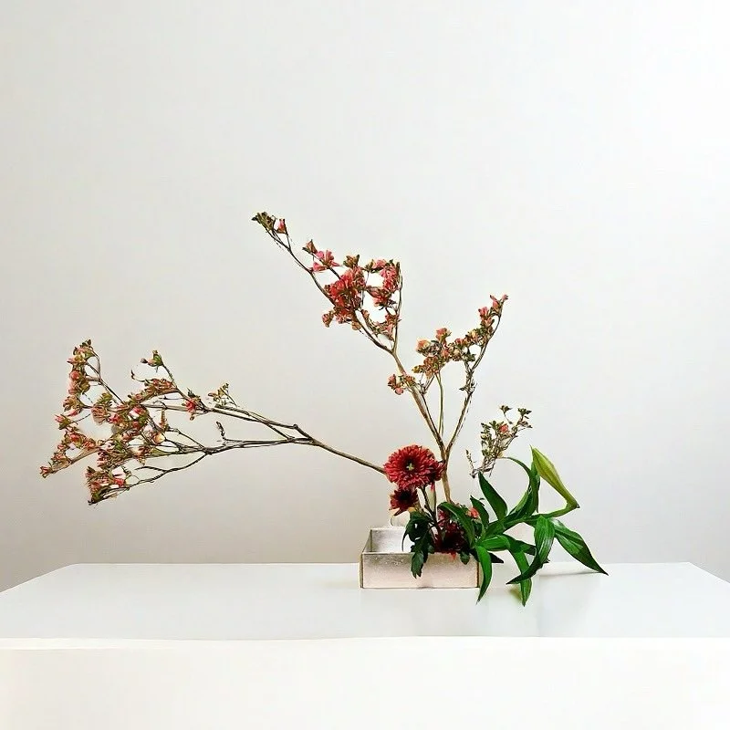 A minimalist flower arrangement in a silver square vase on a white surface, featuring pink blossoms, red flowers, and green leaves against a plain white wall background.
