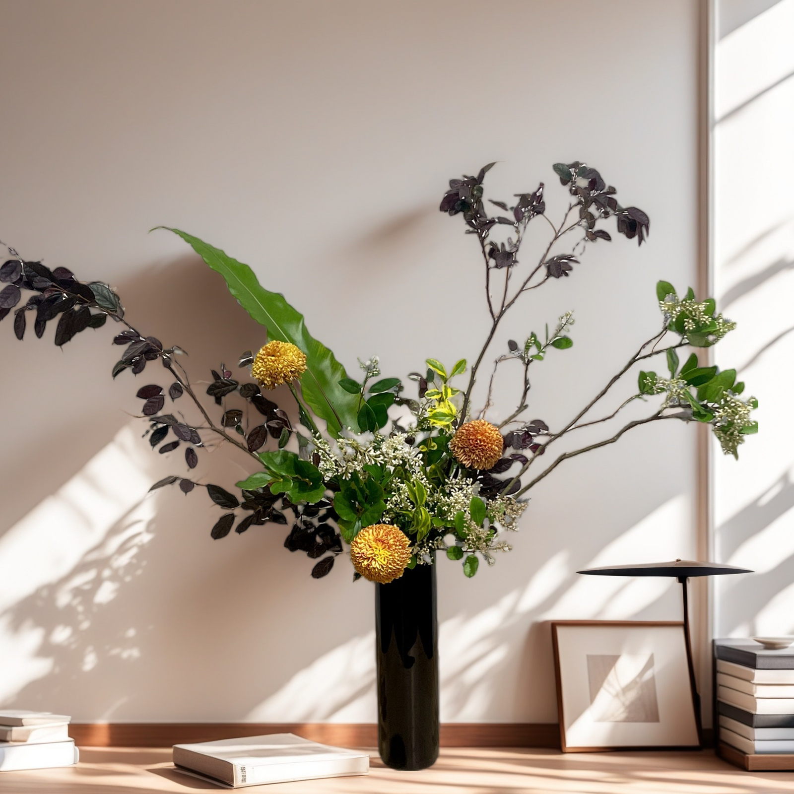 A tall black vase with a floral arrangement on a wooden table. The arrangement includes branches with dark purple leaves, large green leaves, and yellow-orange spherical flowers, with small white flowers and green foliage. A framed artwork and a stack of books are on the right side of the table, with sunlight casting shadows on the wall.