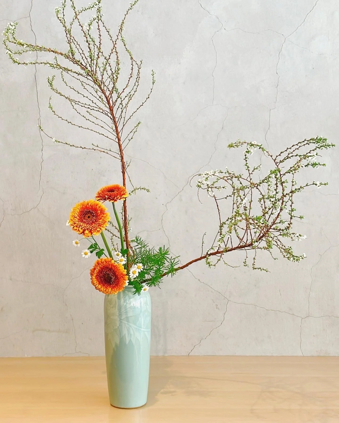 A tall, light-colored ceramic vase with floral patterns containing a mixed arrangement of orange and yellow flowers, small white daisies, and green foliage; some branches are long and sparse with tiny leaves, set against a cracked, off-white wall.