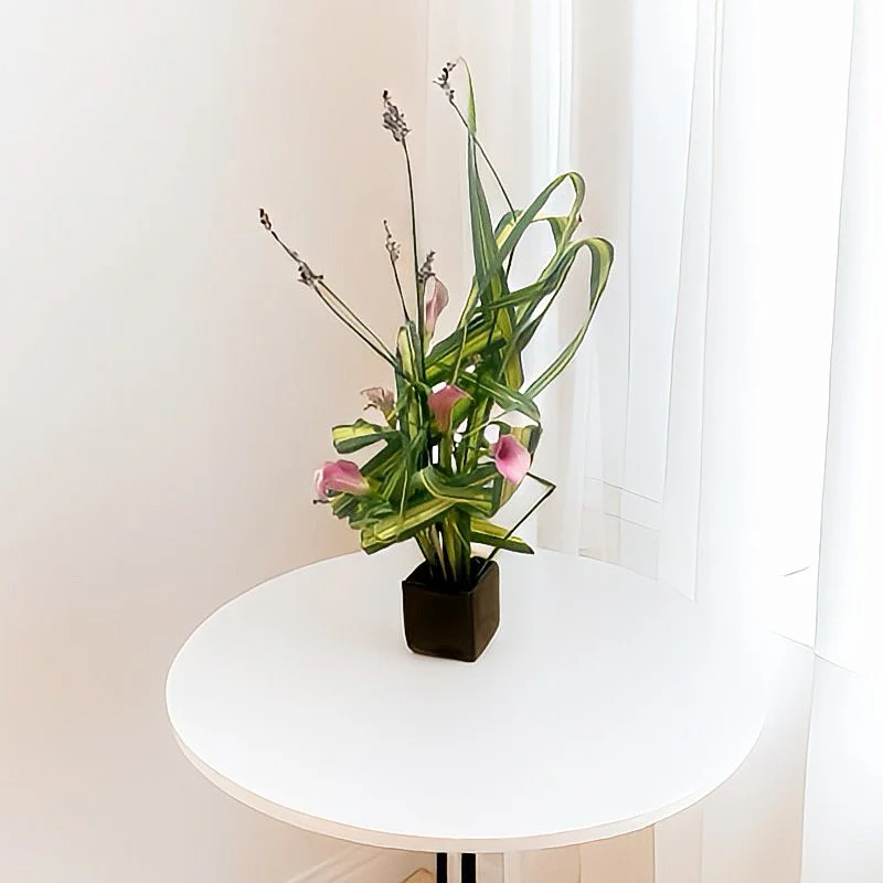 A black square vase with pink and green floral arrangement on a white round table near a sheer white curtain.