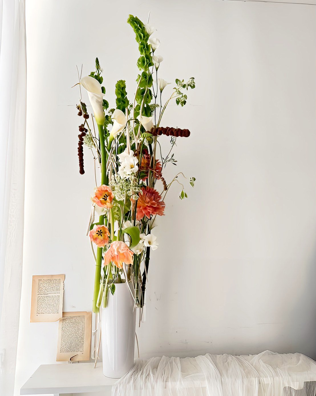 A tall white vase with a large floral arrangement including calla lilies, gladiolas, and various greenery, placed on a small white table next to a white wall with a small framed artwork.