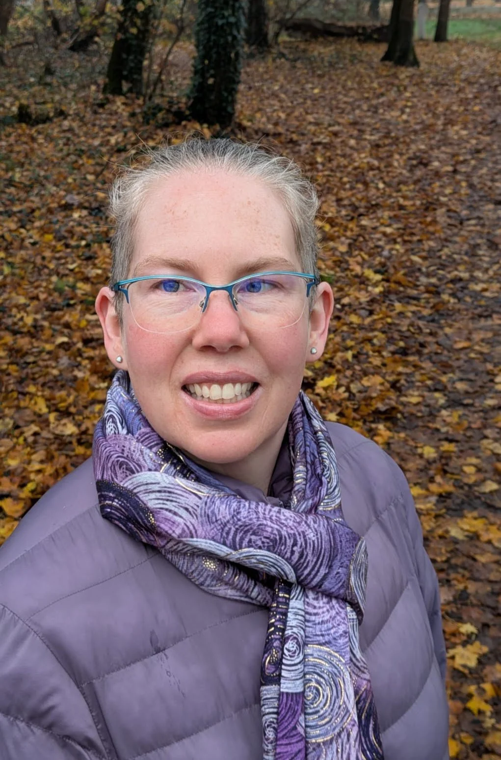 A woman with glasses and earrings taking a selfie outdoors in a park with autumn leaves on the ground and trees in the background.